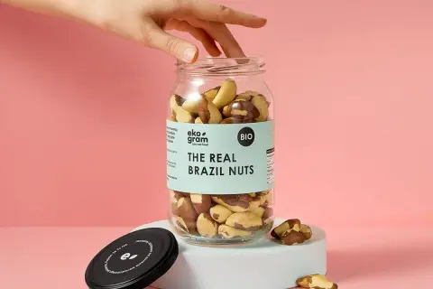 Dłoń sięga po słoik pełen orzechów brazylijskich. Na etykiecie widnieje napis "THE REAL BRAZIL NUTS".