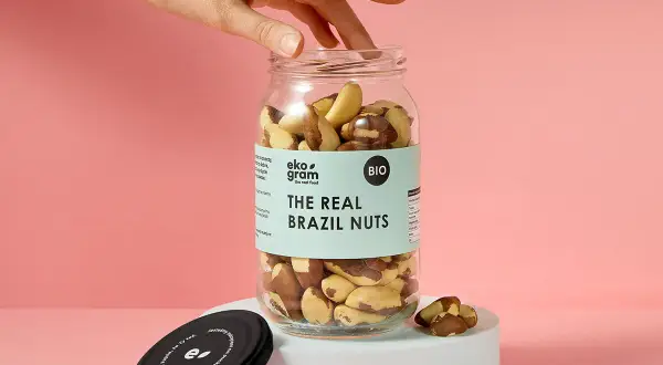 Dłoń sięga po słoik pełen orzechów brazylijskich. Na etykiecie widnieje napis "THE REAL BRAZIL NUTS".