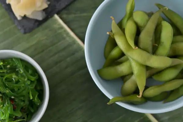 Miseczka z zieloną fasolką edamame, obok sałatka z wodorostów i plasterki imbiru na liściu bananowca.